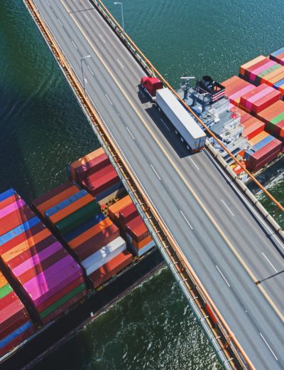 Aerial drone view of a container ship navigating beneath a semi truck crossing a suspension bridge.