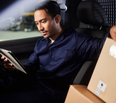 Young male delivery person using a mobile app on a digital tablet while sitting in his van before leaving a shipping and distribution warehouse
