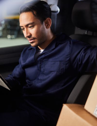 Young male delivery person using a mobile app on a digital tablet while sitting in his van before leaving a shipping and distribution warehouse